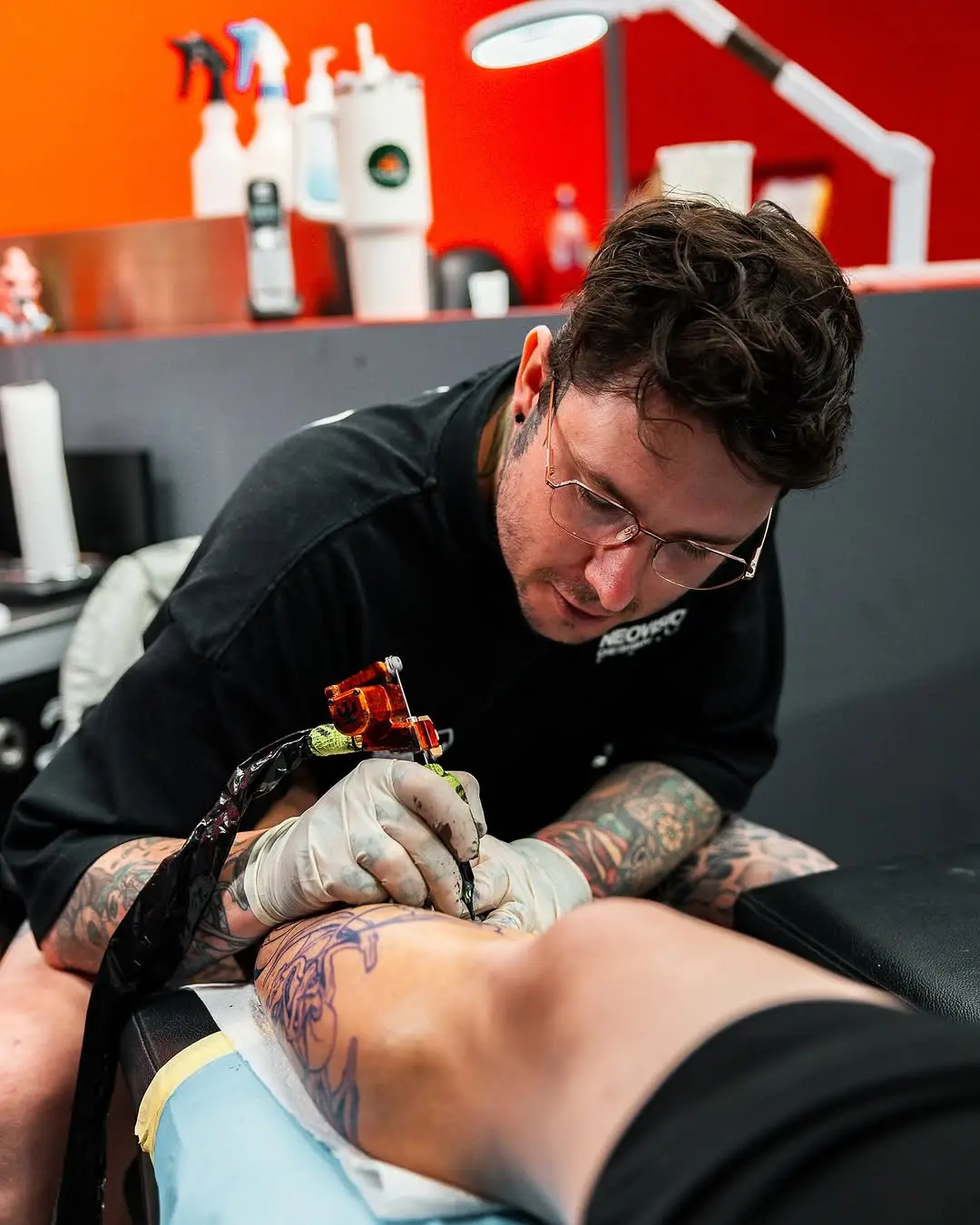 A tattooist leaning in to line a forearm piece, working from a stencil already applied to the skin