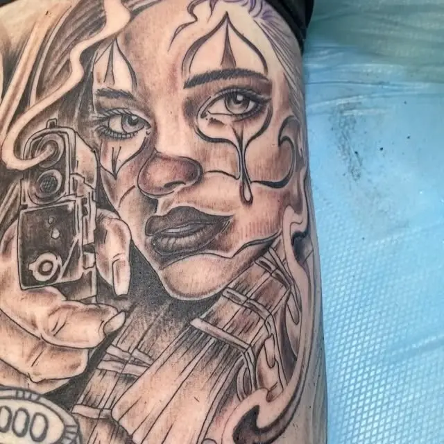 A black and grey Chicano style portrait of a woman with teardrop markings, holding a tattoo machine