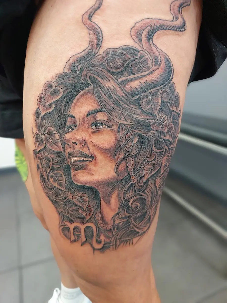 A black and grey Medusa portrait on a thigh, serpents rising from her hair and a Scorpio glyph beneath