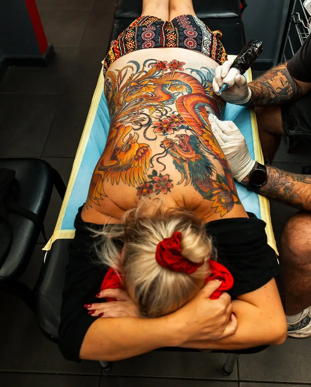 A full colour phoenix and dragon back piece in progress, photographed from above while the artist works on the lower back
